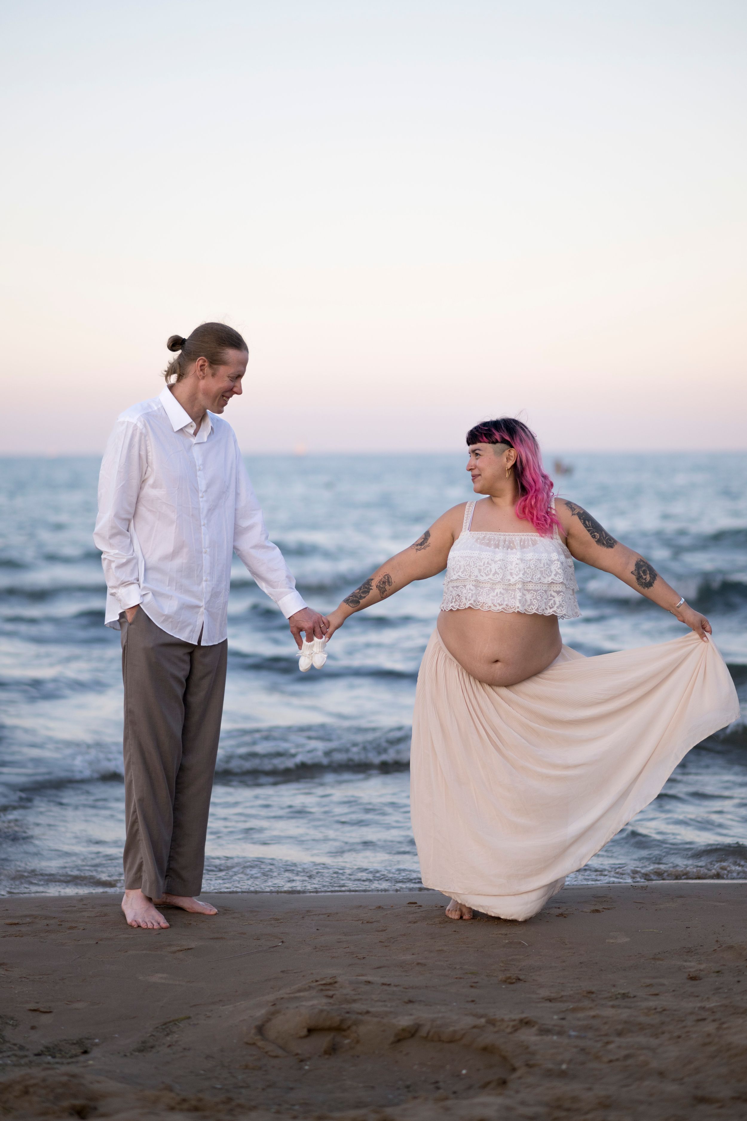 A pregnant couple hold a tiny pair of baby booties in between them. They stand by the water at the beach, him in slacks, her in a skirt.