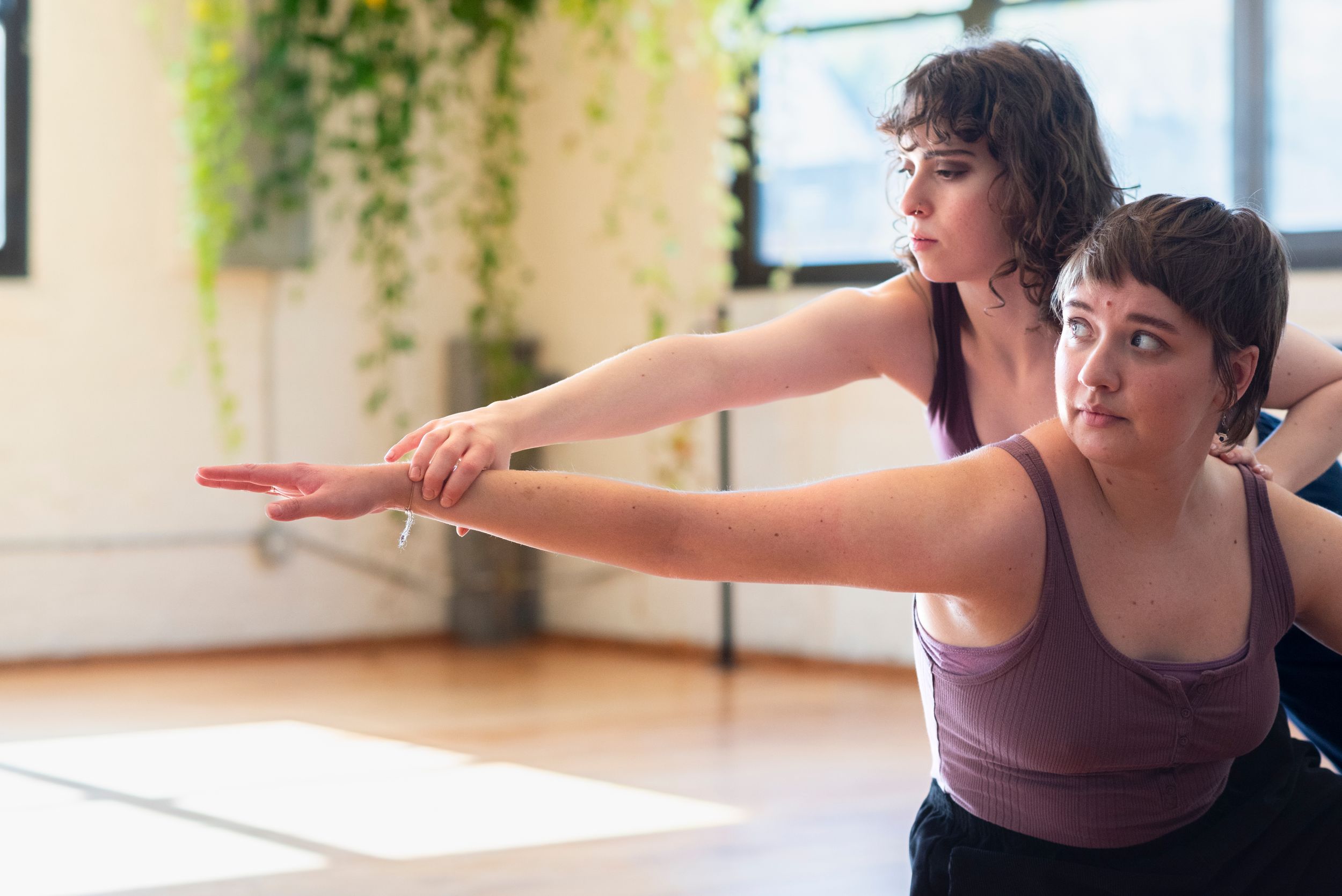 Two dancers perform, one holding the wrist and arm of the other as they reach together, in a sun-soaked dance studio.
