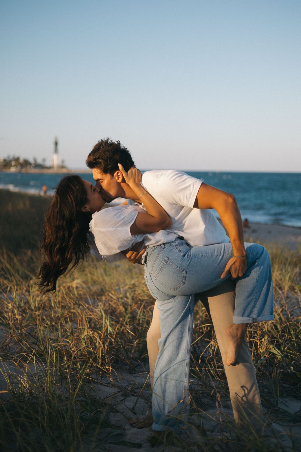 couple kissing in front of beach and lighthouse in background