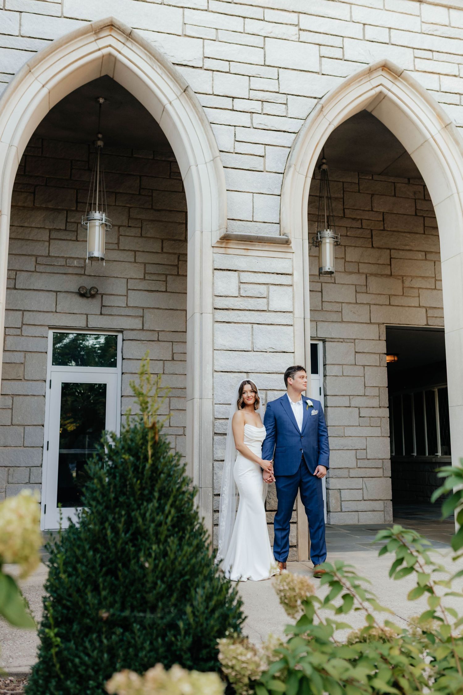 Wedding couple poses under stone archways of elegant church building.