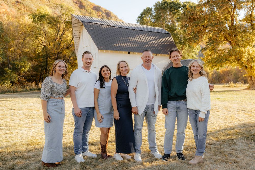 Family of seven smiling together outdoors near a white barn during autumn.