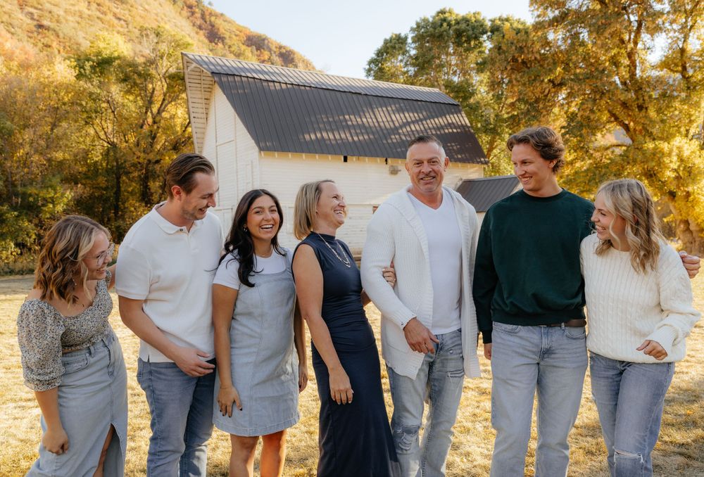 Laughing family group of seven posing casually near a white barn in fall.