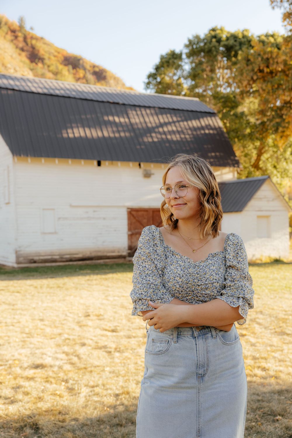 Smiling blonde woman in floral top and jeans standing outdoors near a white barn.