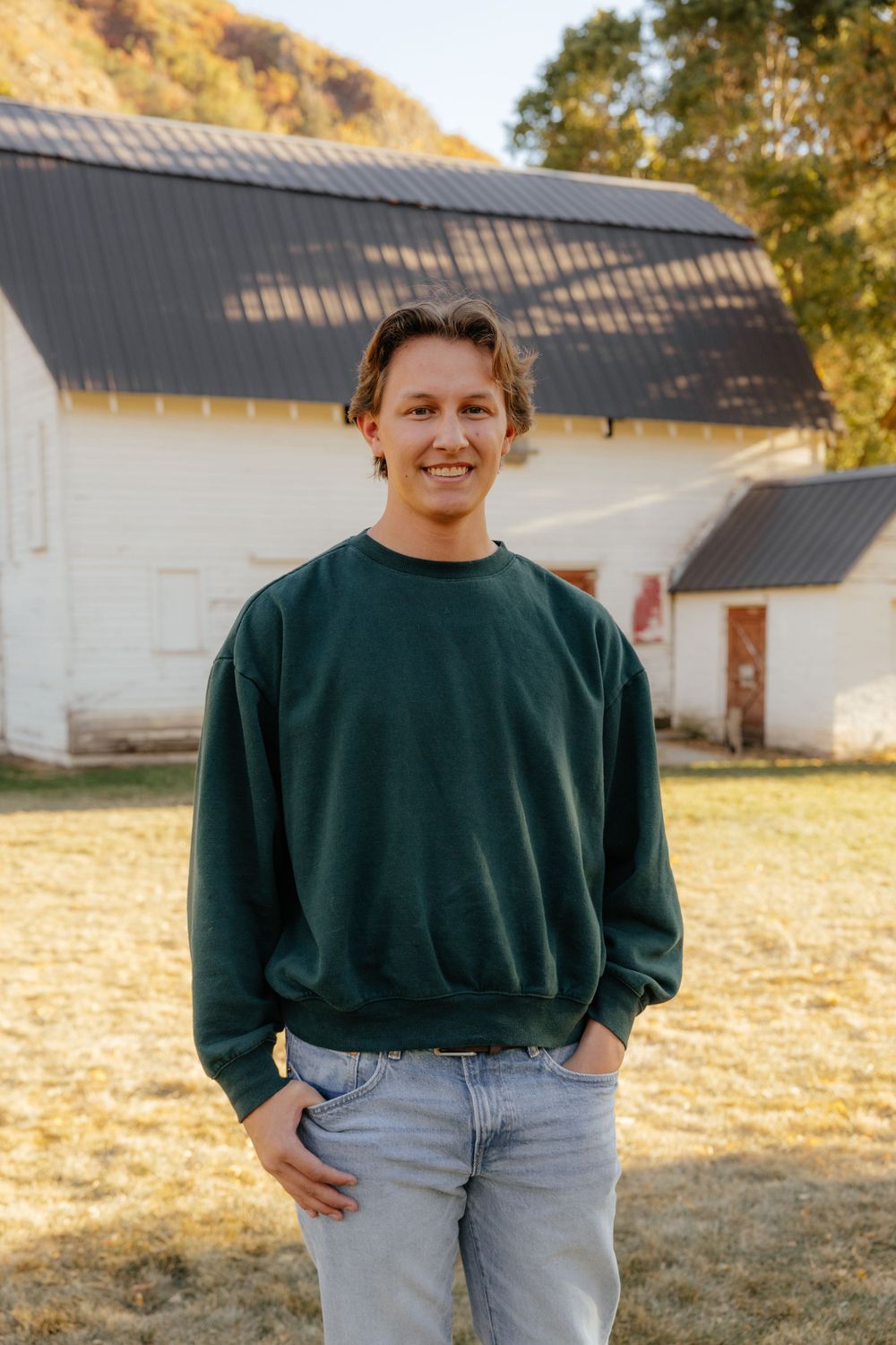 Smiling young man in green sweatshirt standing outdoors near a white barn in fall.
