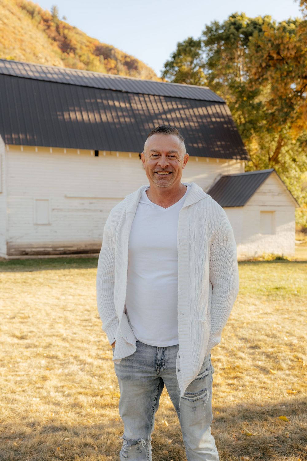 Smiling middle-aged man in white cardigan standing outdoors near a white barn.