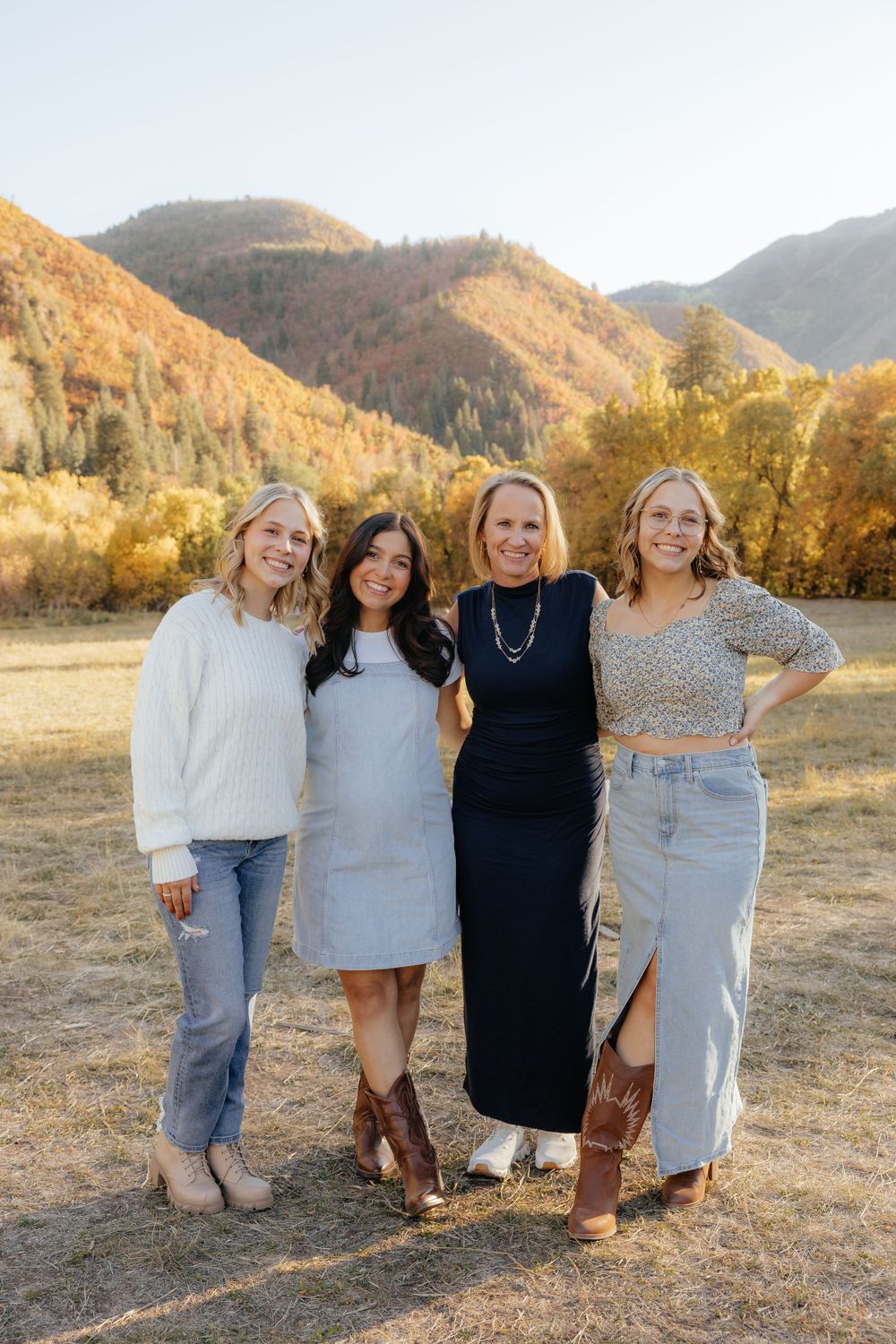 Four women smiling together in a sunny autumn mountain meadow, wearing casual fall outfits including jeans and skirts.