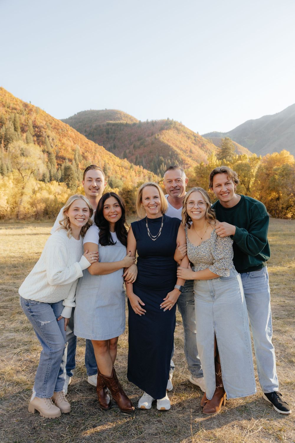 Family of seven smiling together in an open field with colorful autumn mountains in the background.