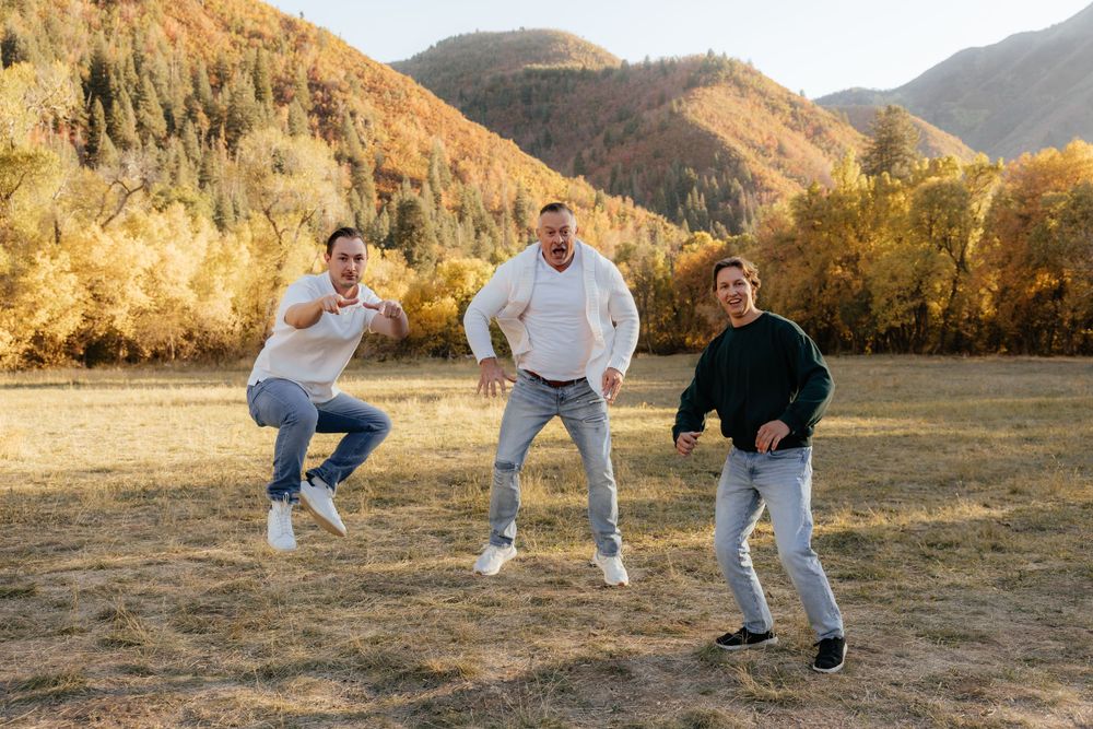 Three men jumping and laughing playfully in an open field with colorful autumn mountains behind them.