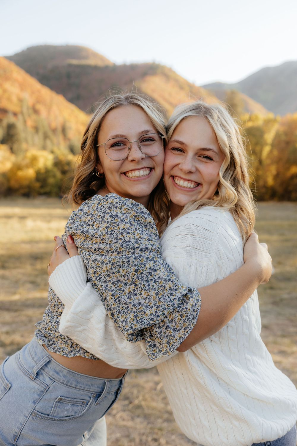 Two blonde women hugging and smiling in a warm autumn mountain meadow during a family photo session.