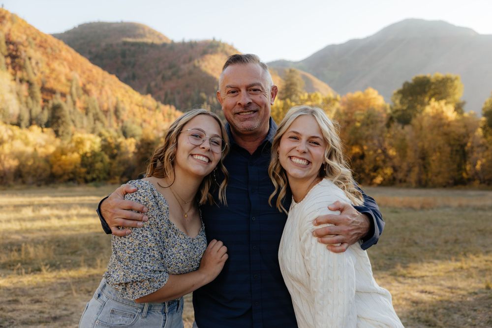 A man with arms around two blonde women smiling together in a golden autumn mountain meadow.