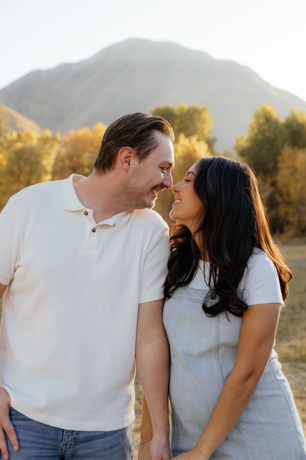 A couple nearly touching noses and smiling at each other in a sunny autumn mountain landscape.