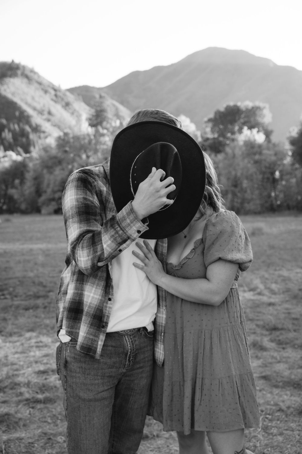 Couple kissing behind a cowboy hat in a black and white mountain field photo.