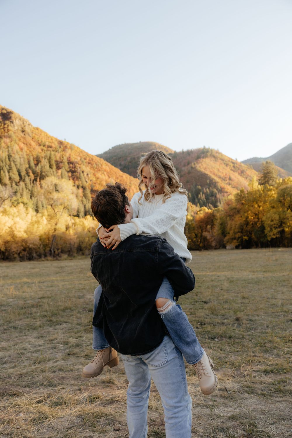 Man lifting smiling woman in white sweater in a golden autumn mountain meadow at sunset.