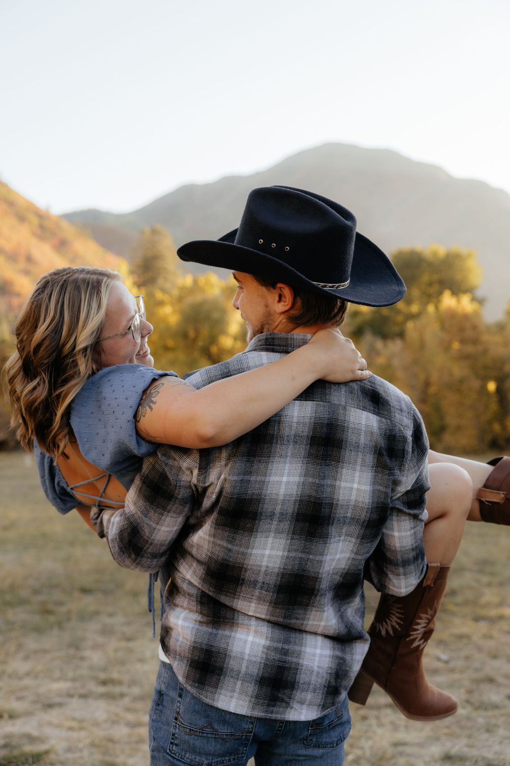Man in cowboy hat carrying woman in his arms in a sunny autumn mountain field.