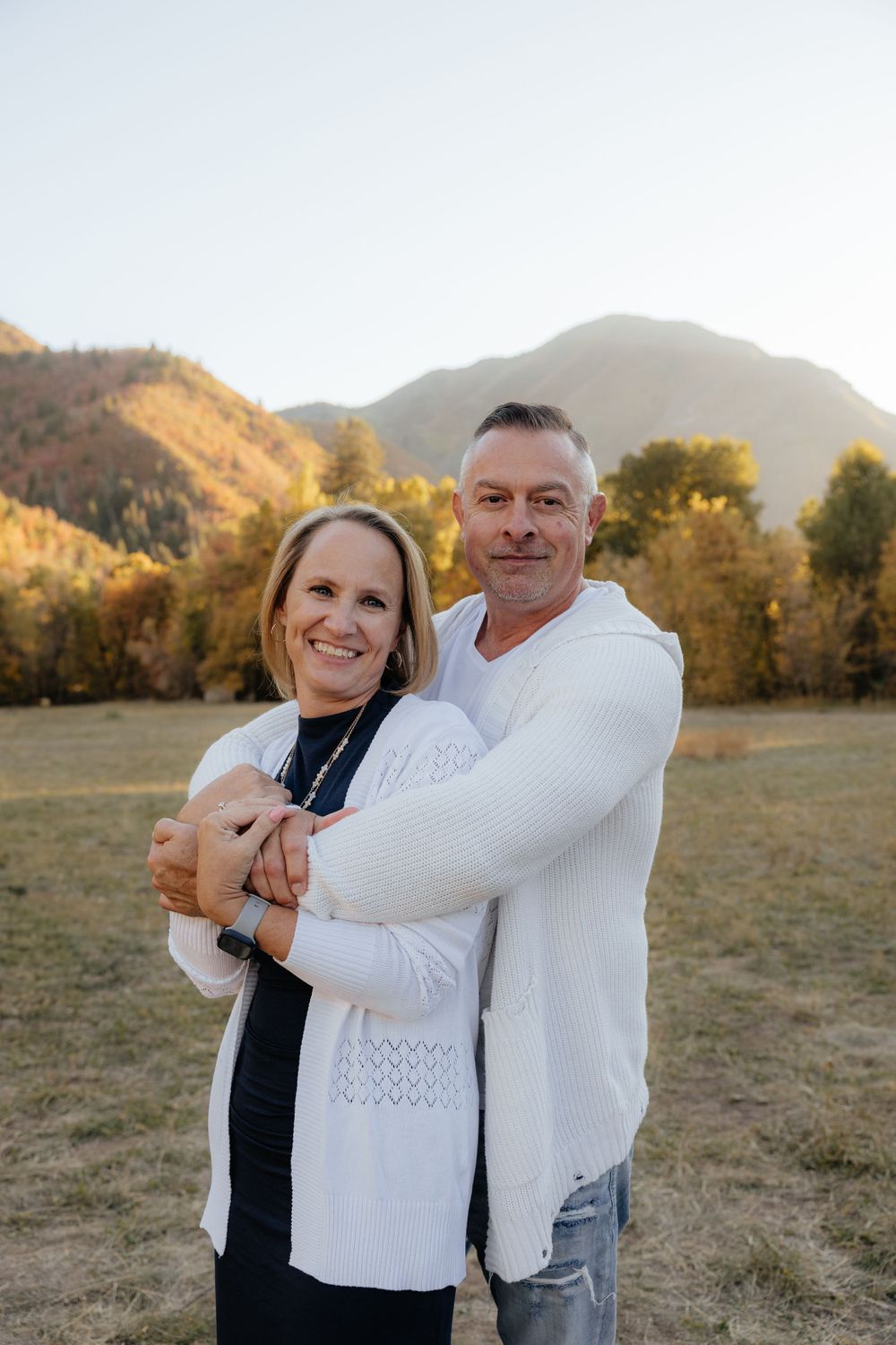 Smiling couple embracing in a field with colorful autumn mountains and trees in background.