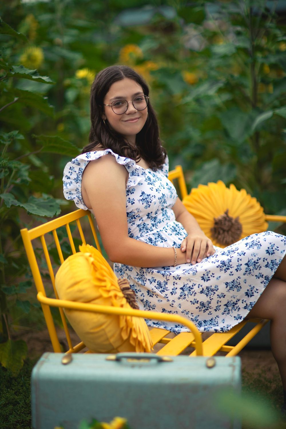Person seated on yellow chair in sunflower garden wearing floral dress.