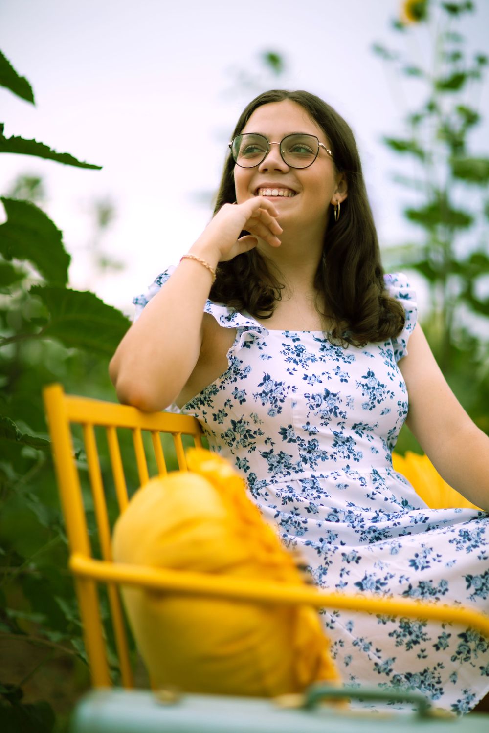 A summer portrait in a floral blue and white dress sitting in a yellow chair among garden foliage.