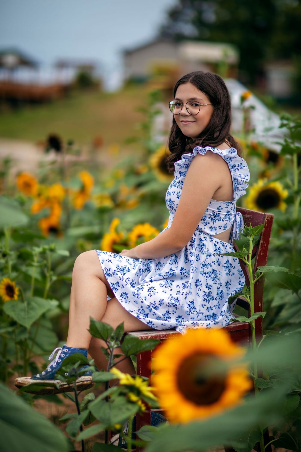A portrait amidst vibrant sunflowers wearing a blue floral dress and glasses in a garden setting.