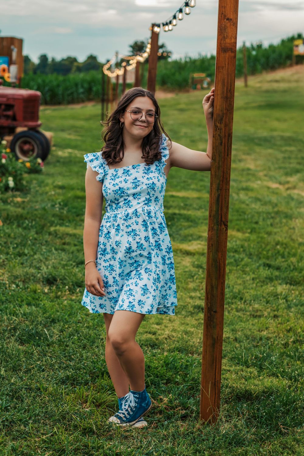A person in a blue floral summer dress and sneakers poses by a wooden post in a grassy field.