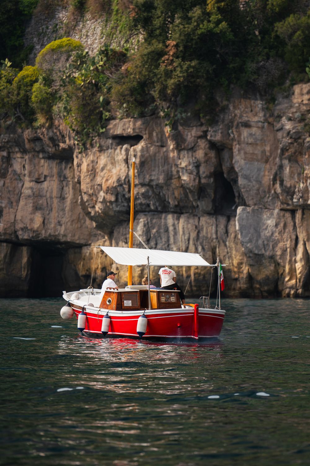 Red and white boat sailing near dramatic limestone cliffs along the Amalfi Coast.