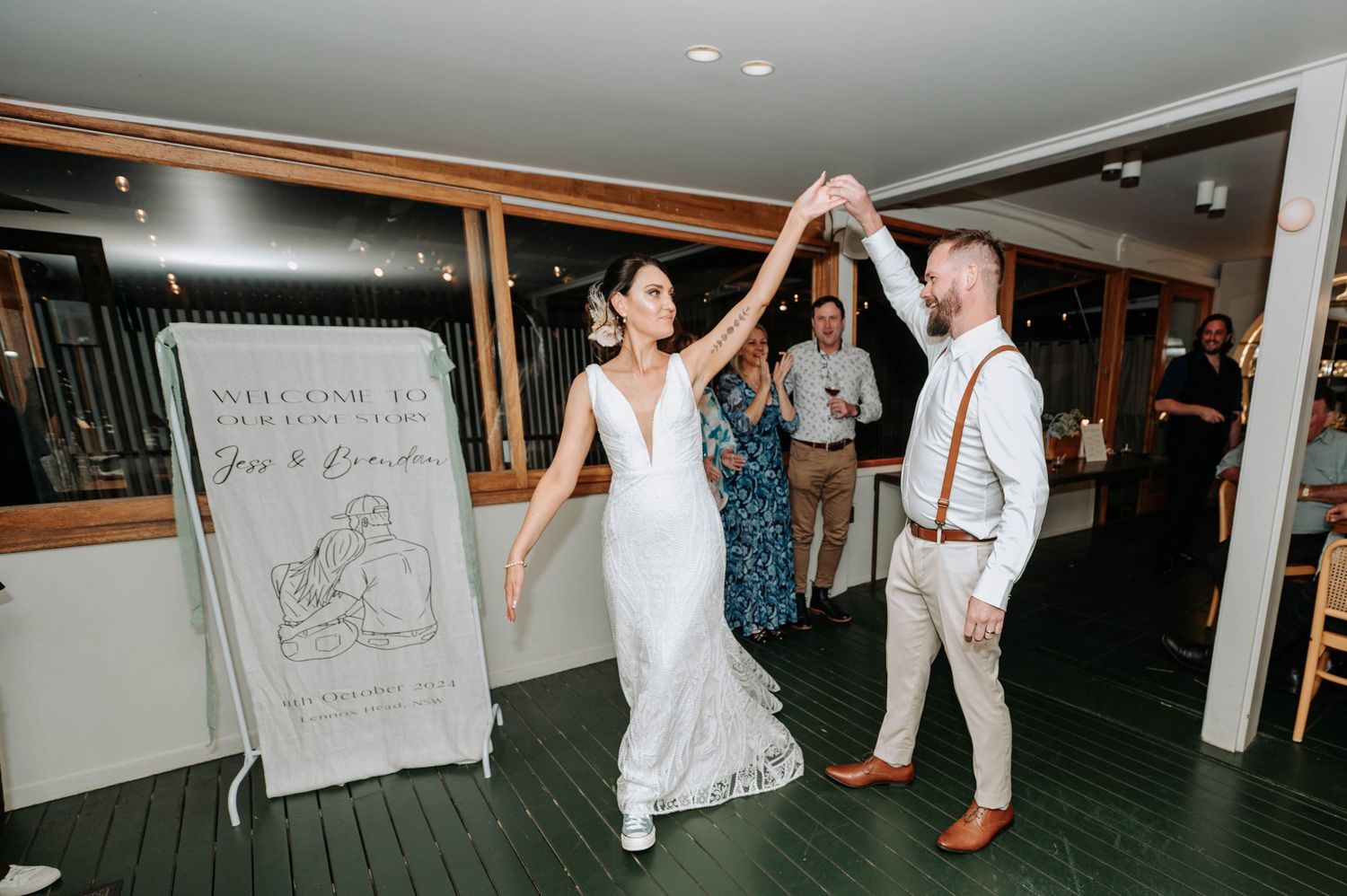 Wedding couple dancing together on indoor wooden dance floor with welcome sign visible.