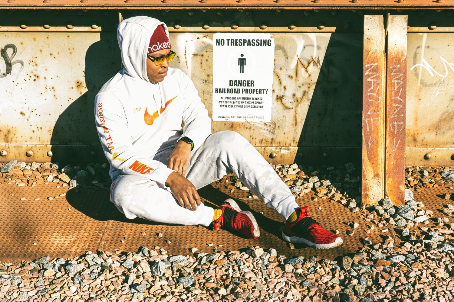 Person in white Nike hoodie and pants sits casually against weathered wall in sunlight.
