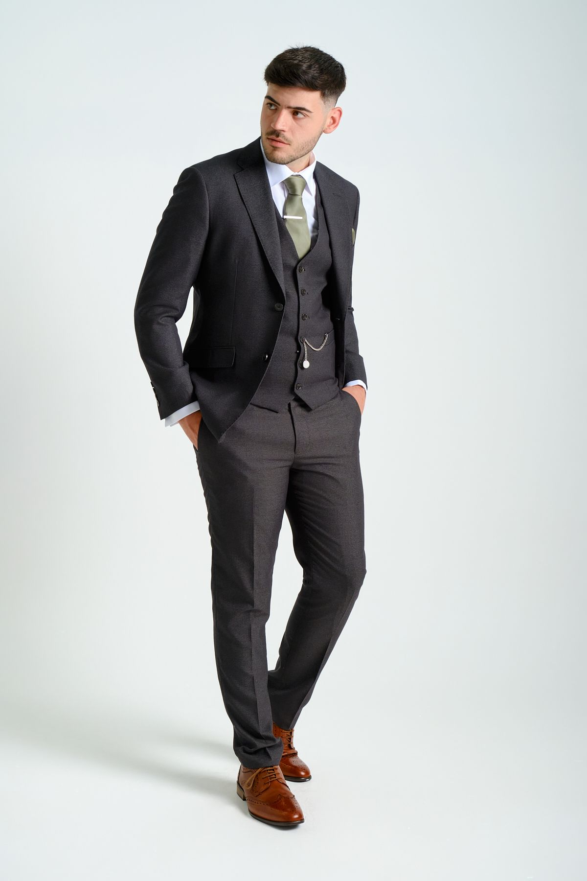 Man in a dark suit and green tie, posing in a classic style with hands in pockets, looking to the side