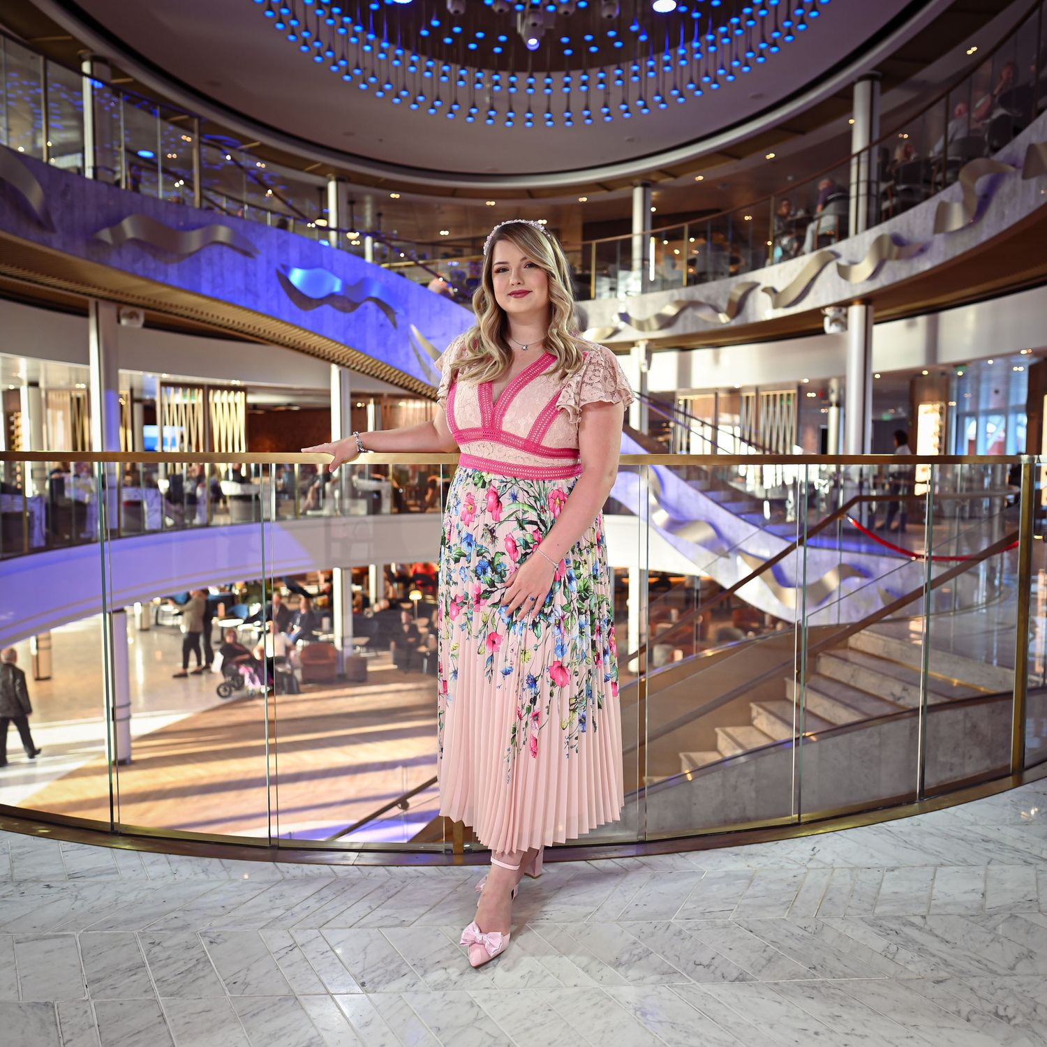 Woman in floral dress poses on elegant cruise ship staircase with blue lighting.