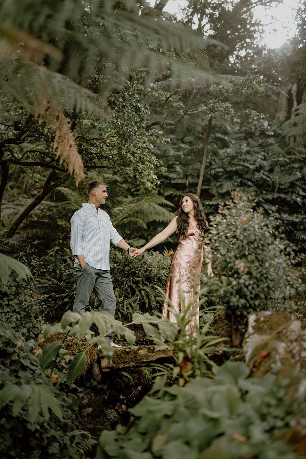 Couple holds hands while exploring a lush garden path surrounded by green ferns and foliage.