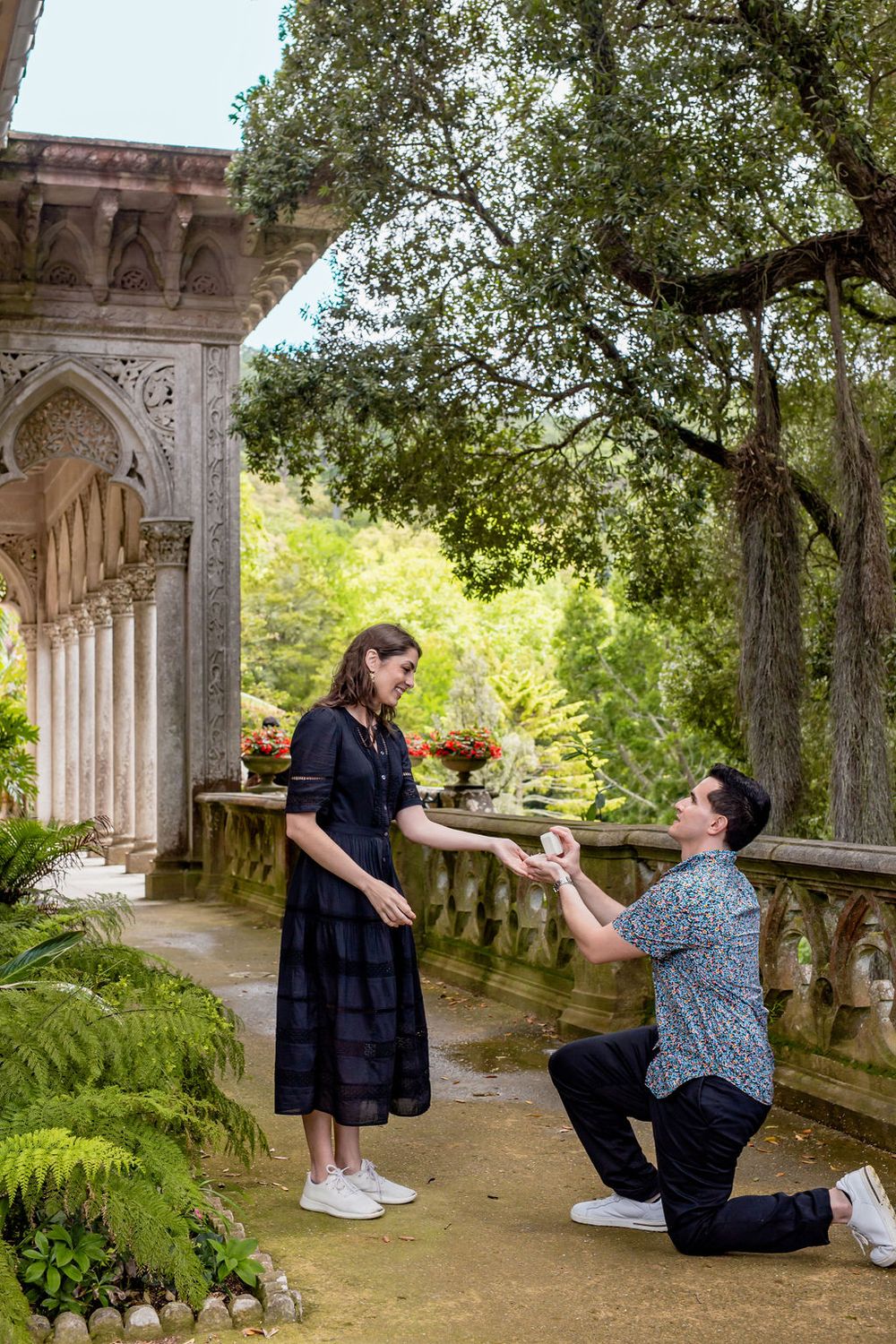 A couple shares a romantic proposal moment in a scenic garden colonnade with stone pillars and lush greenery.