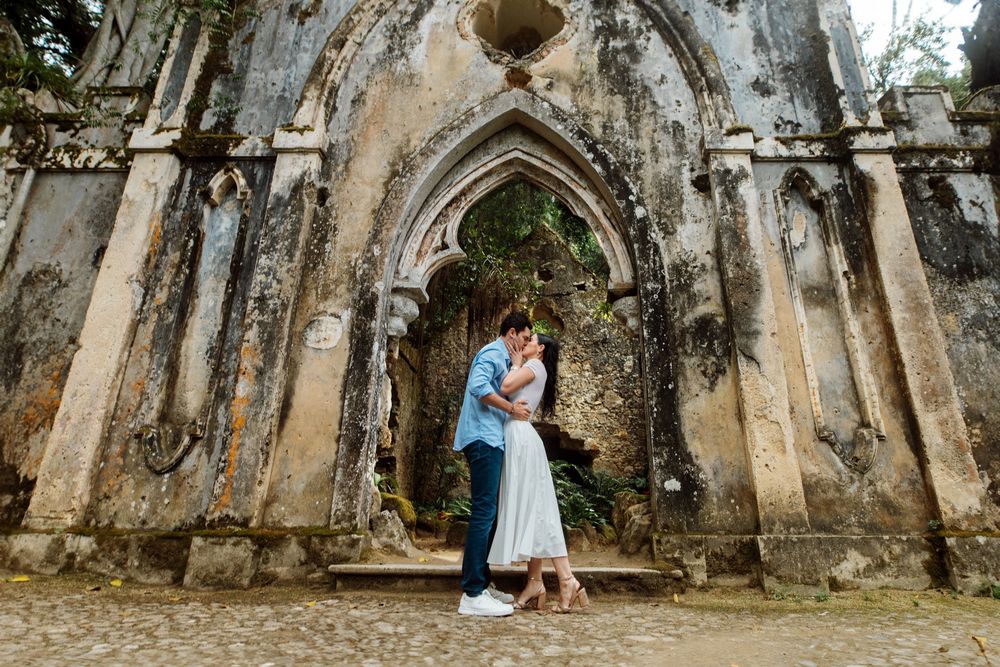 A couple embraces in front of a weathered Gothic stone archway with ornate architectural details.