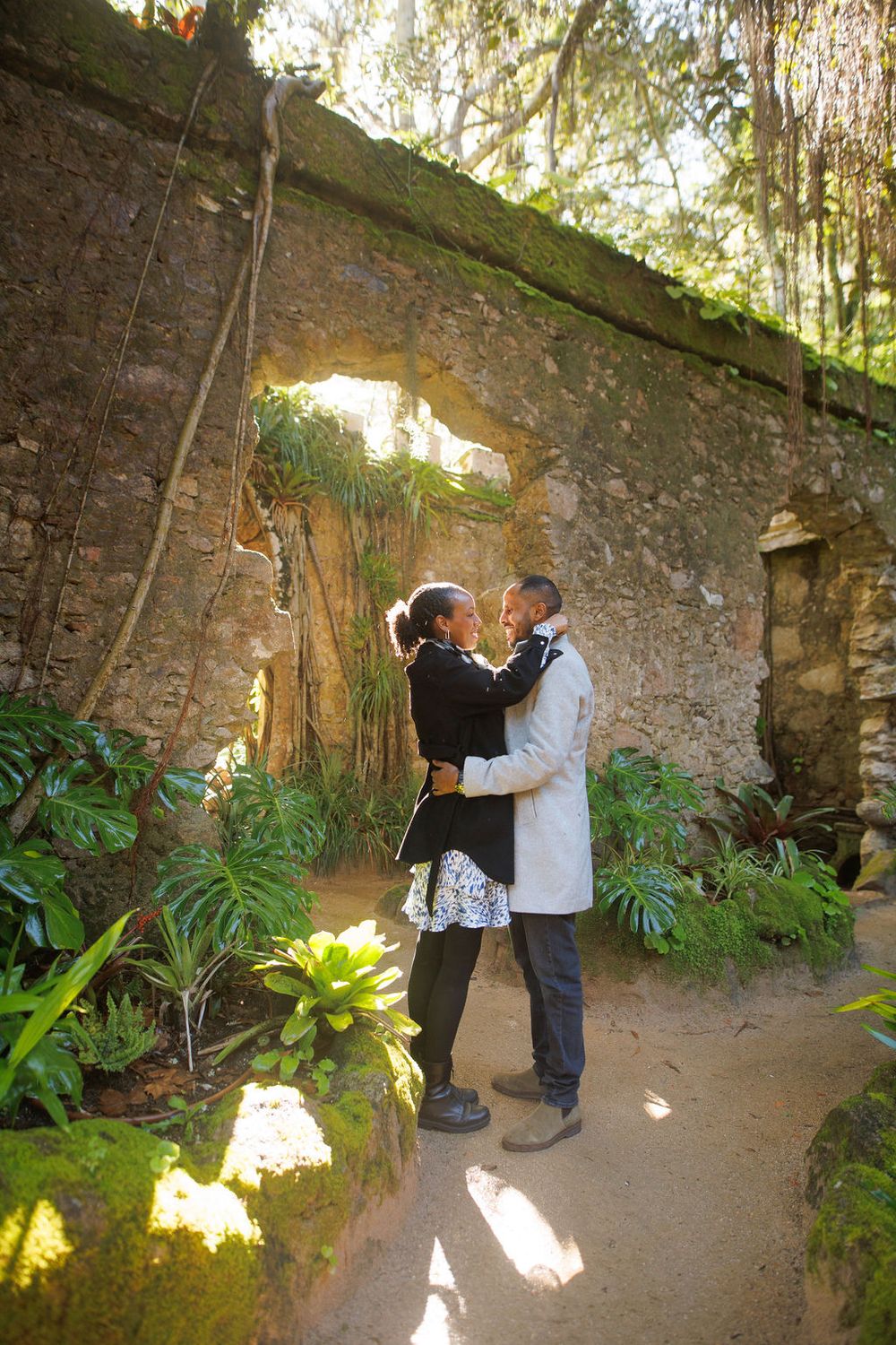 A romantic moment captured in a secluded garden grotto with stone walls and tropical plants creating a magical atmosphere.