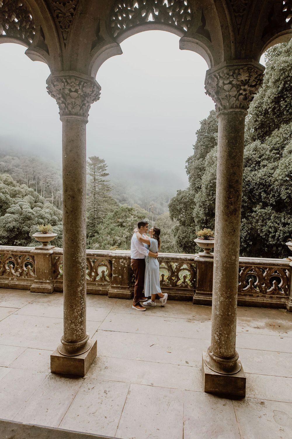 A romantic moment captured through stone archways on a misty terrace at a historic palace or castle.