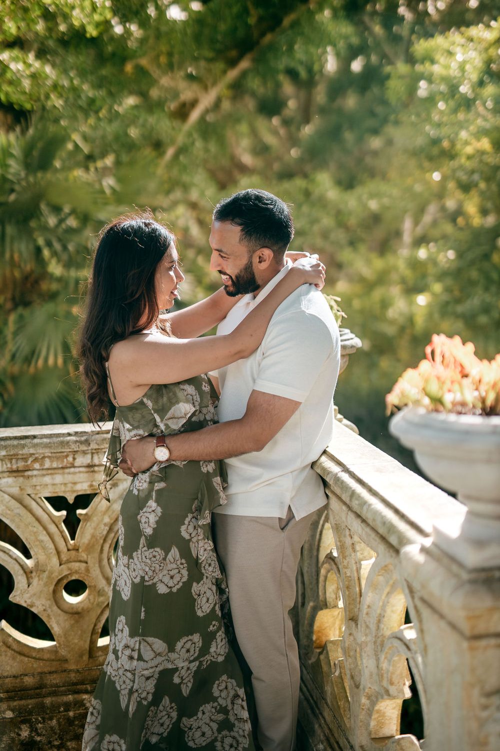 Romantic couple embraces on ornate stone balcony surrounded by lush greenery in natural sunlight.