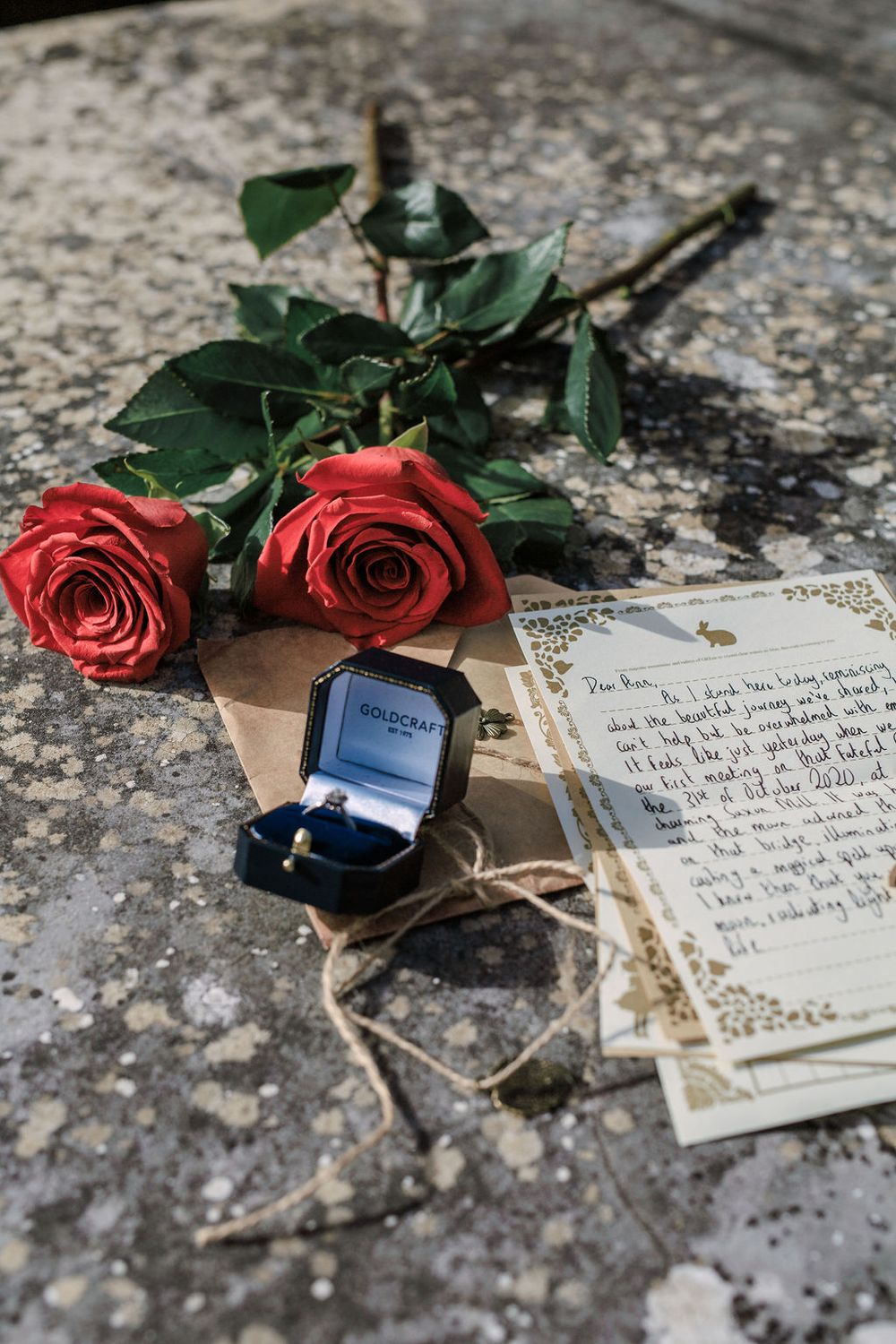 Red roses lay beside handwritten letters and an engagement ring on a textured stone surface in a romantic proposal setup.