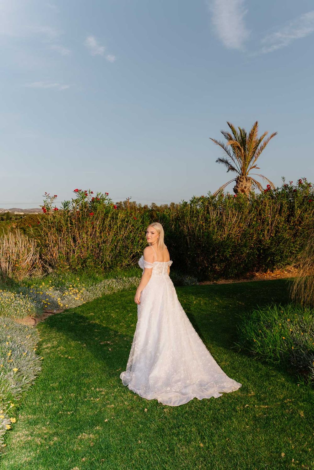 A person in a flowing white wedding dress walks through a lush garden path with palm trees and flowers in the background.