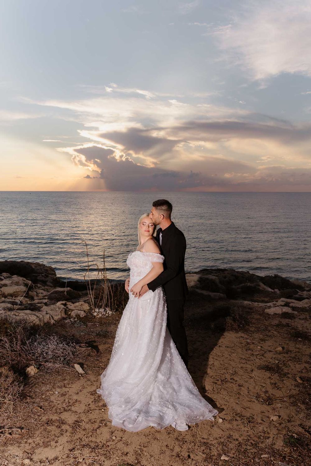 A couple shares a romantic moment at sunset on a rocky coastal cliff overlooking the ocean.
