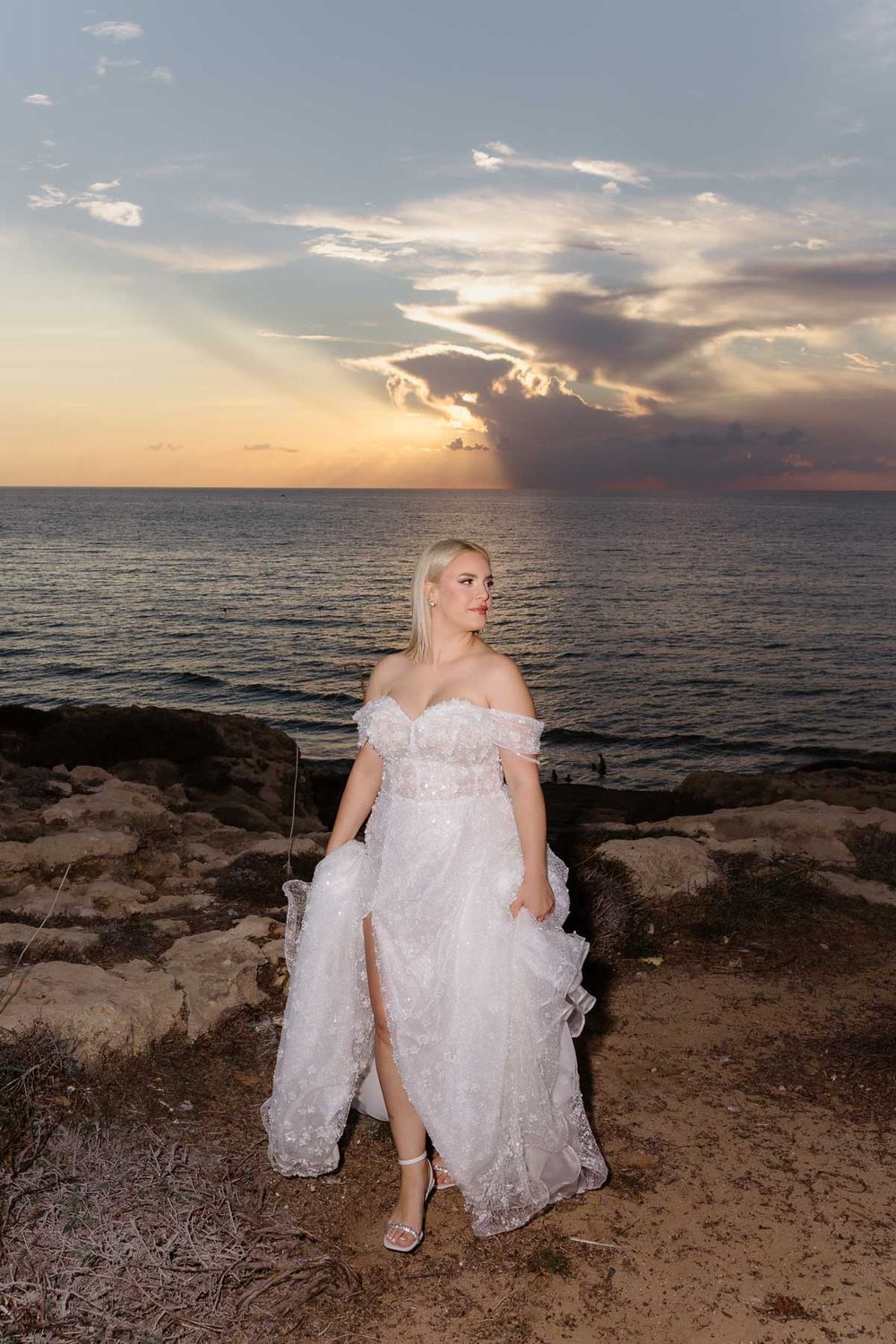 A scenic coastal sunset photoshoot features someone in a flowing white lace wedding dress sitting on rocky cliffs by the ocean.