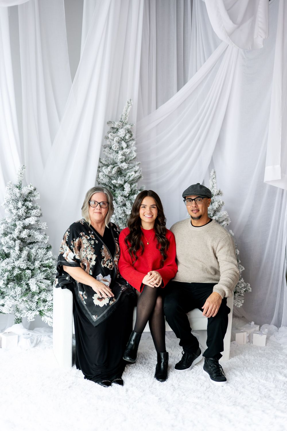 Family of three sitting and posing together surrounded by frosted Christmas trees and white decor.