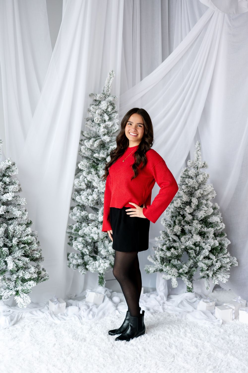 Smiling young woman in red sweater and black skirt poses confidently in winter holiday setting.