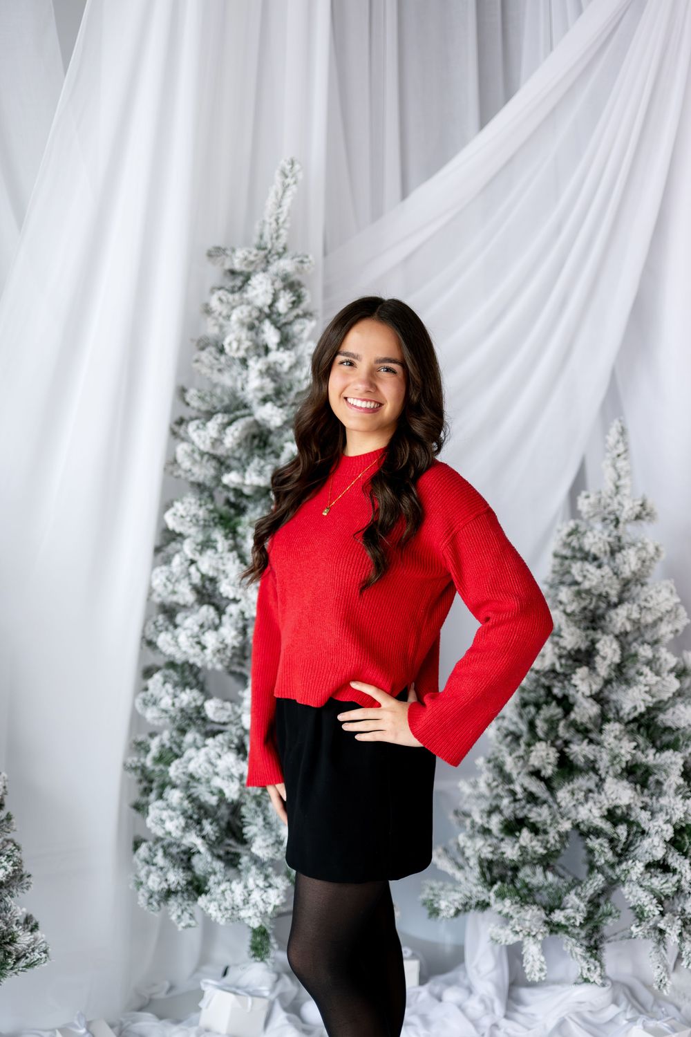 Young woman in red top and black skirt smiles beside snow-flocked Christmas trees in studio.