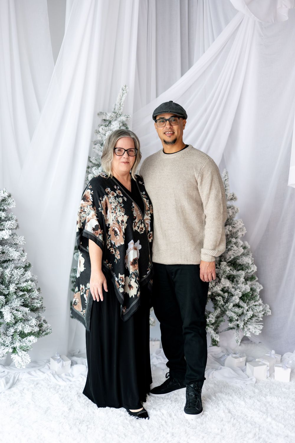 Couple posing together in front of flocked Christmas trees and elegant white curtain backdrop.