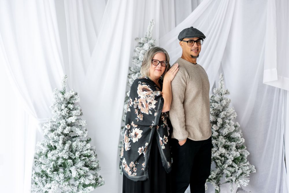 Couple posing back to back in front of flocked Christmas trees and white draped backdrop.