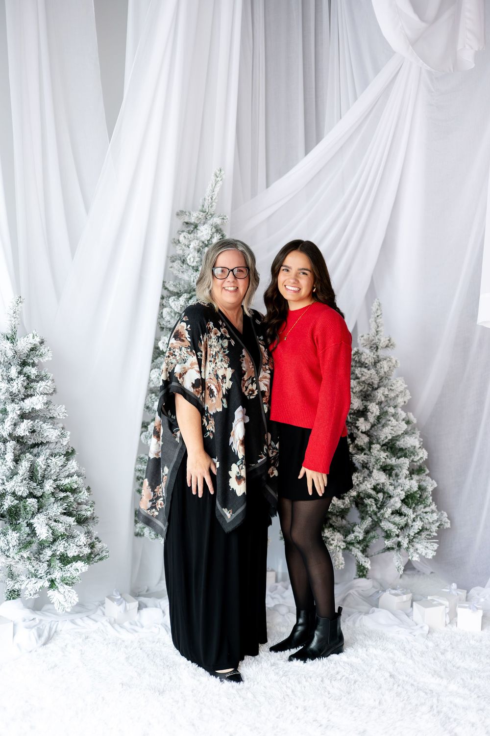 Two women smiling together in festive outfits before snow-covered Christmas trees in studio.