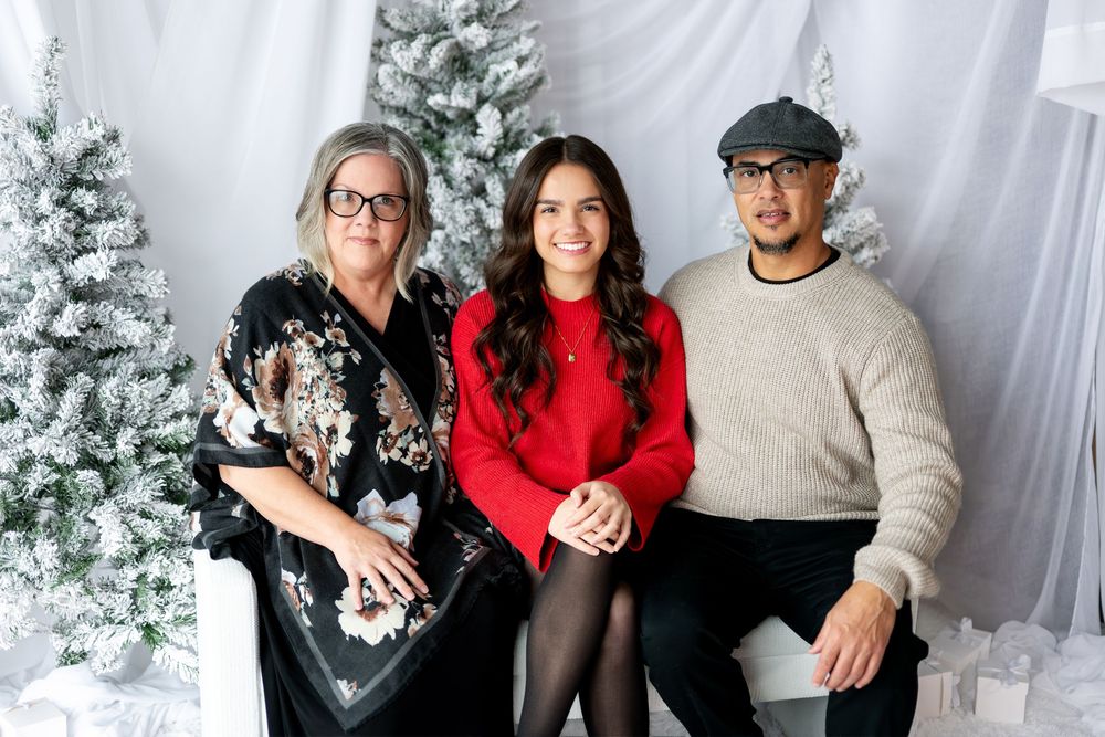 Family of three poses together in front of snowy Christmas trees, woman in red sweater flanked by parents.