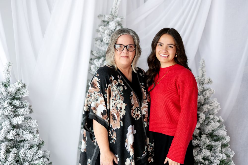 Two women posing together in front of flocked Christmas trees and white curtain backdrop.