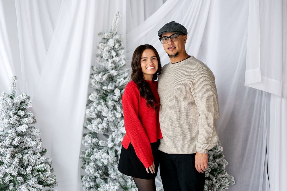 Young couple in coordinating sweaters posing before snow-flocked Christmas trees in studio.