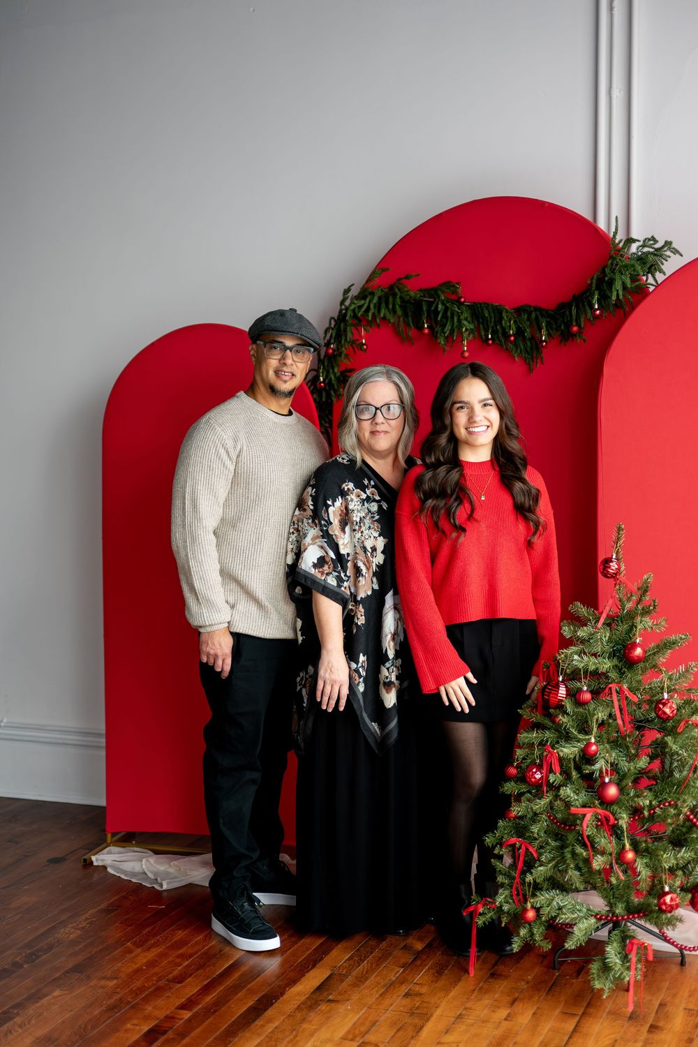 Three people pose for a festive Christmas photo in front of red arch backdrop with holiday decorations and Christmas tree.