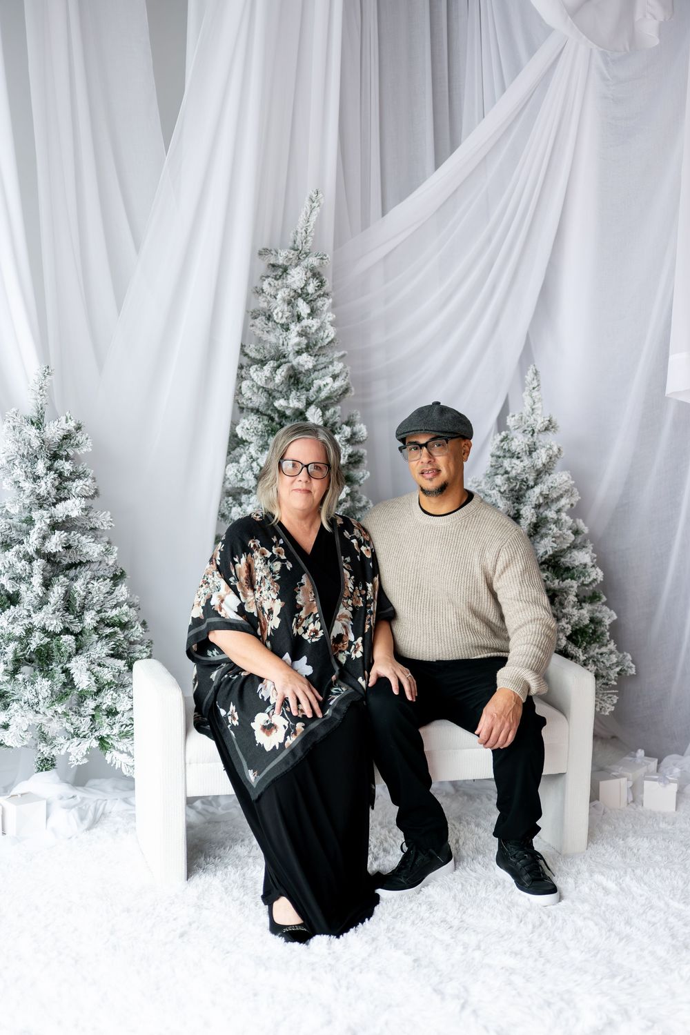 Couple in holiday attire sits together in front of snow-flocked Christmas trees and white drapes.
