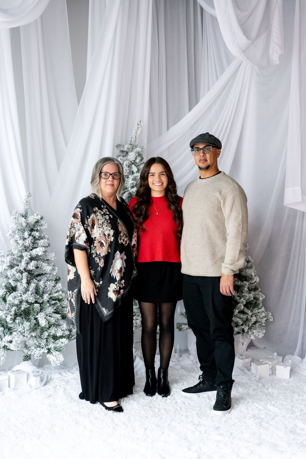 Family of three stands together smiling in festive outfits before snow-covered Christmas trees.